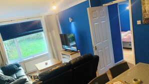 Un salon avec un canapé en cuir noir, un téléviseur posé sur un meuble, une table à manger avec des chaises, et une porte donnant sur une chambre.