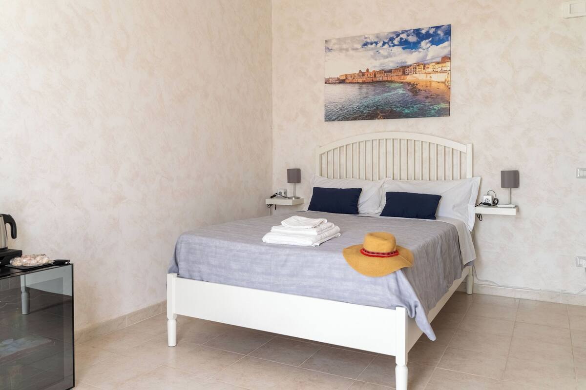 A neatly made bed with a gray bedspread, two pillows, and a folded towel. A hat is placed on the bed. There is a painting of a coastal scene on the wall.