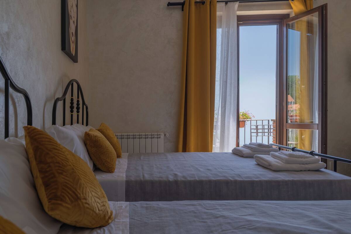 A hotel room with two beds, a window with yellow curtains, and a view of the outside.