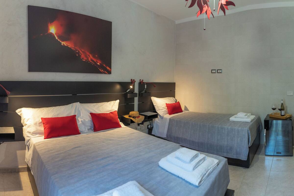 A hotel room with two beds, a nightstand, a lamp, a painting of a volcano, and a minibar.