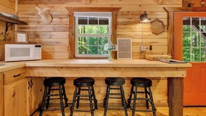 Interior de una cabaña de madera con una zona de cocina que incluye un microondas, una ventana y un conjunto de taburetes de bar.