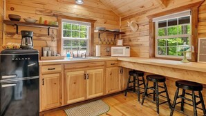 Cocina compacta con armarios de madera, un refrigerador Galanz, microondas y un lavamanos. Hay tres taburetes y una ventana con vista a la vegetación.