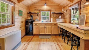 Interior de una cabaña con paredes de madera, una cocina con refrigerador, estufa y taburetes de bar.