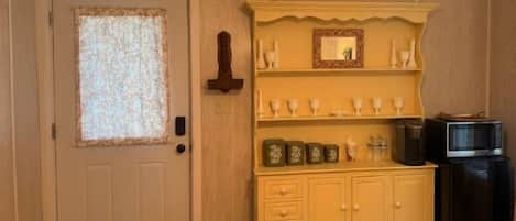 Una habitación con una puerta, una estantería de madera con objetos decorativos, un mueble pequeño y un televisor.