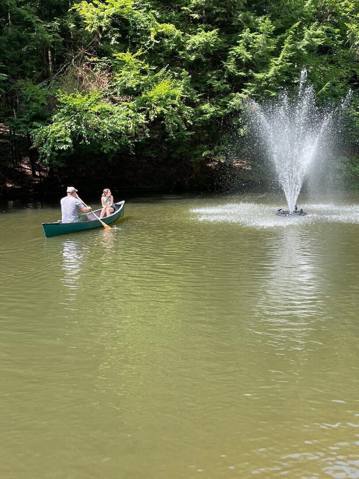 Um casal em um barco canoa verde em um lago calmo com uma fonte ao fundo.
