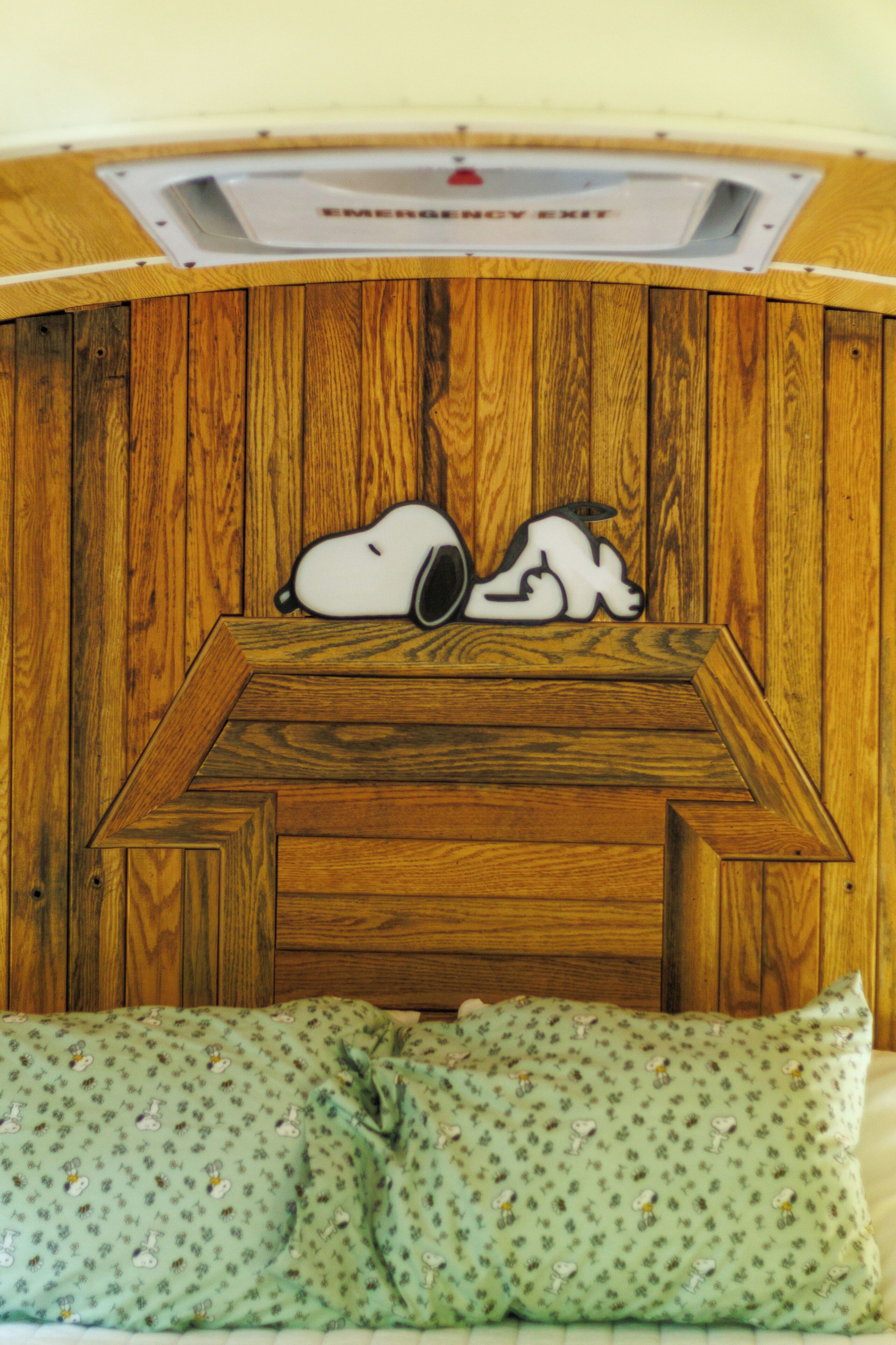 A room with a wooden wall, a bed with a green pillow, and a ceiling with an emergency exit sign.