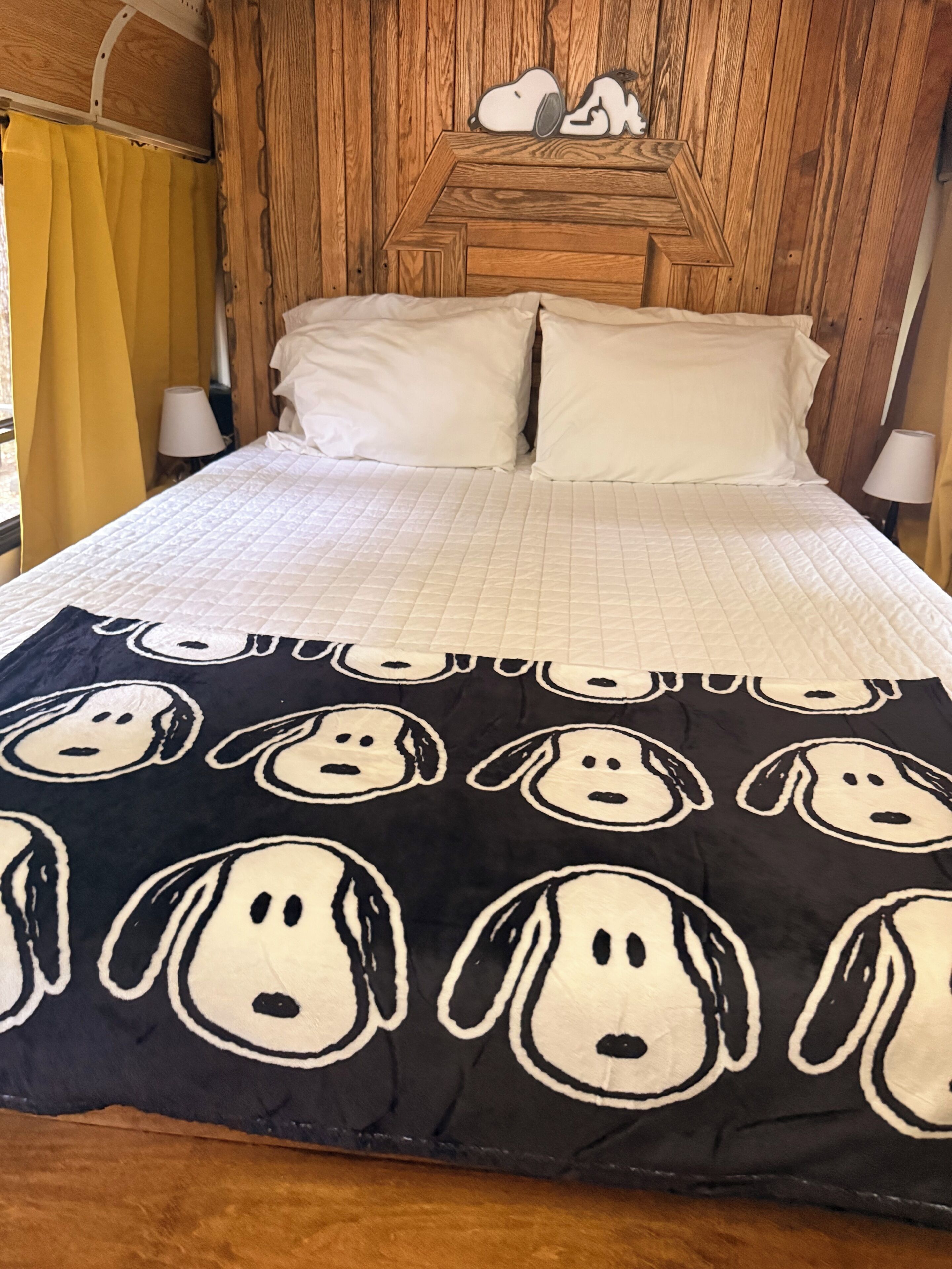 Un lit recouvert d’une couette blanche matelassée, une couverture noire ornée de motifs de chiens de dessins animés et une tête de lit en bois décorée avec un motif de Snoopy.