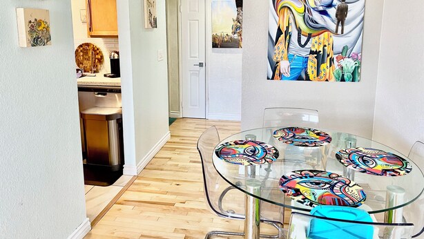 A modern dining area with a glass table and chairs, a kitchen in the background, and a colorful mural on the wall. - Artsy home near Downtown and Balboa Park (San Diego)