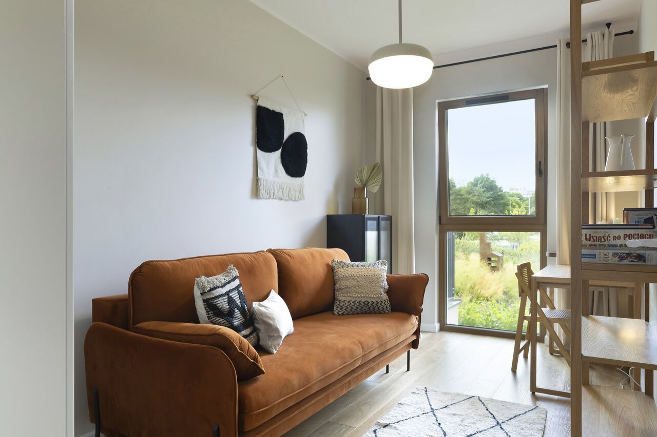 A modern living room with a brown sofa, a dining table, and a large window offering a view of greenery.