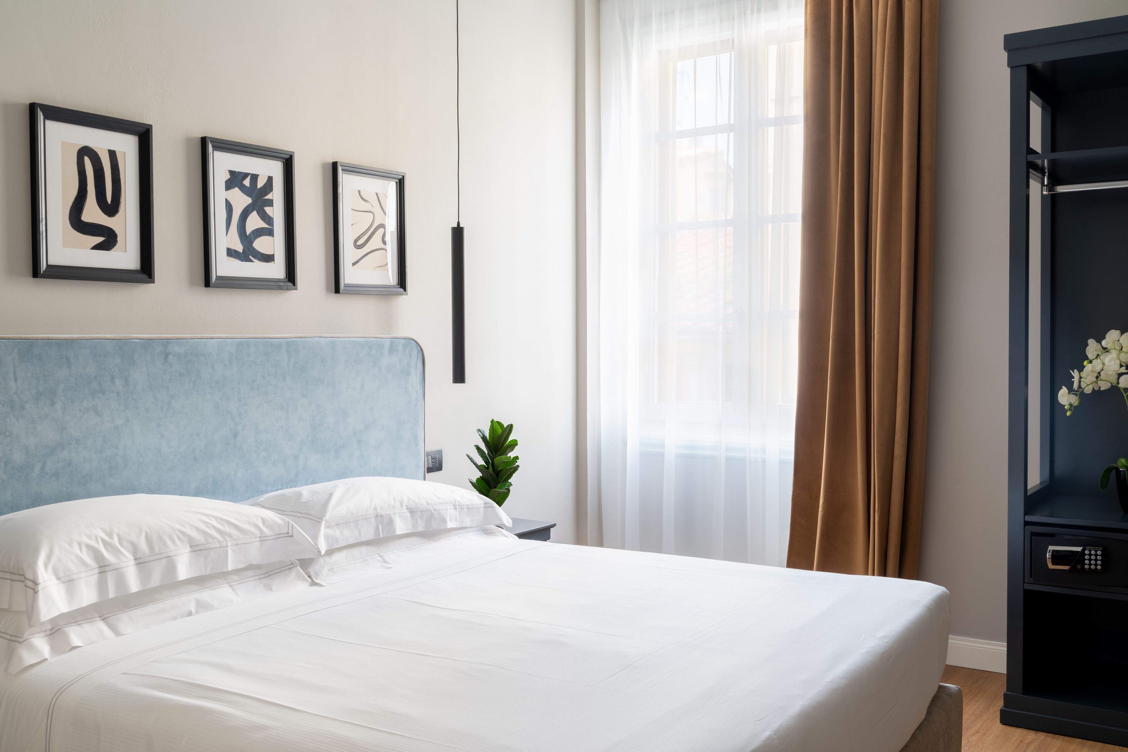 Un letto ben rifatto con lenzuola bianche, una testiera blu e tre opere d'arte astratte incorniciate appese al muro.