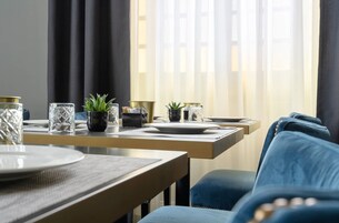 Zona pranzo con tavolo apparecchiato per due, dal design moderno che combina vetro e metalli, due piante in vaso e un divano blu imbottito sullo sfondo.