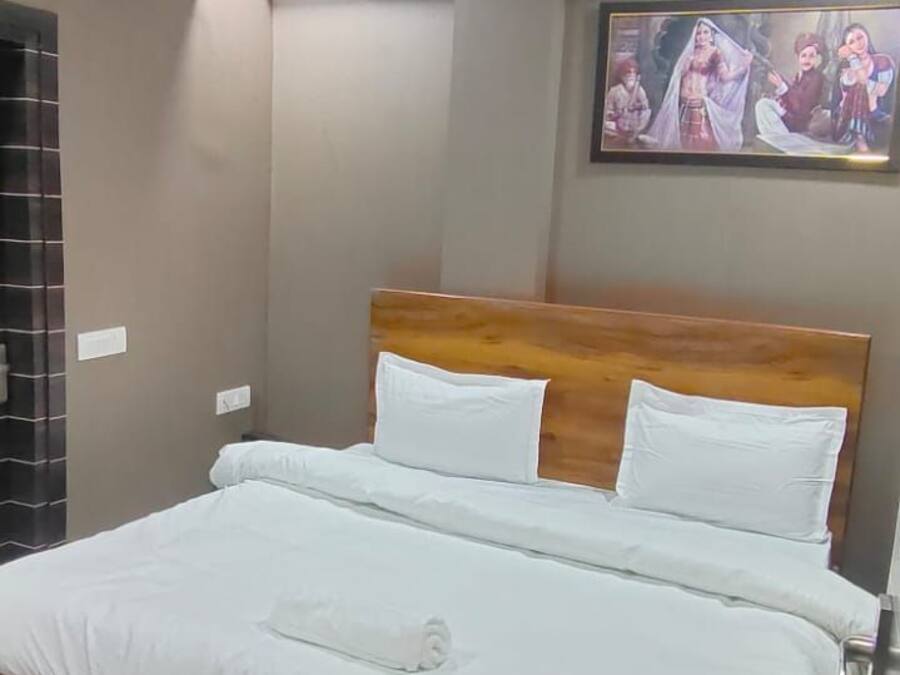 Habitación de hotel con cama de madera, ropa de cama blanca, ventilador de techo, equipo de aire acondicionado, un cuadro en la pared y alfombra estampada.