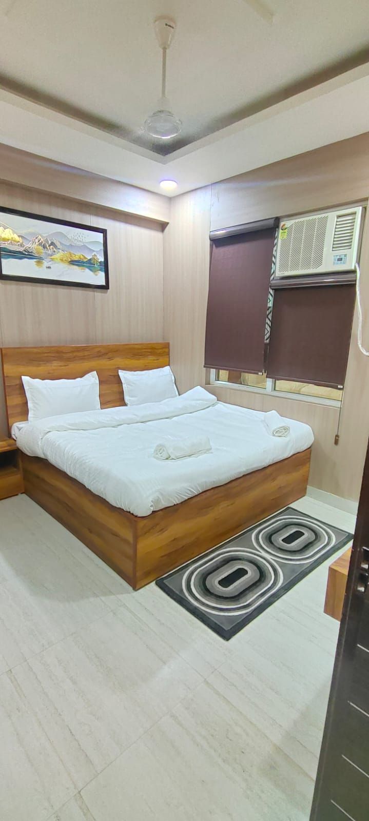 A bedroom with a wooden bed, white bedding, a painting on the wall, an air conditioning unit, and a window with blinds.