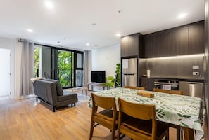 A modern kitchen with dark cabinets, a dining table with chairs, a sofa, and a TV in a living area with large windows and a balcony.