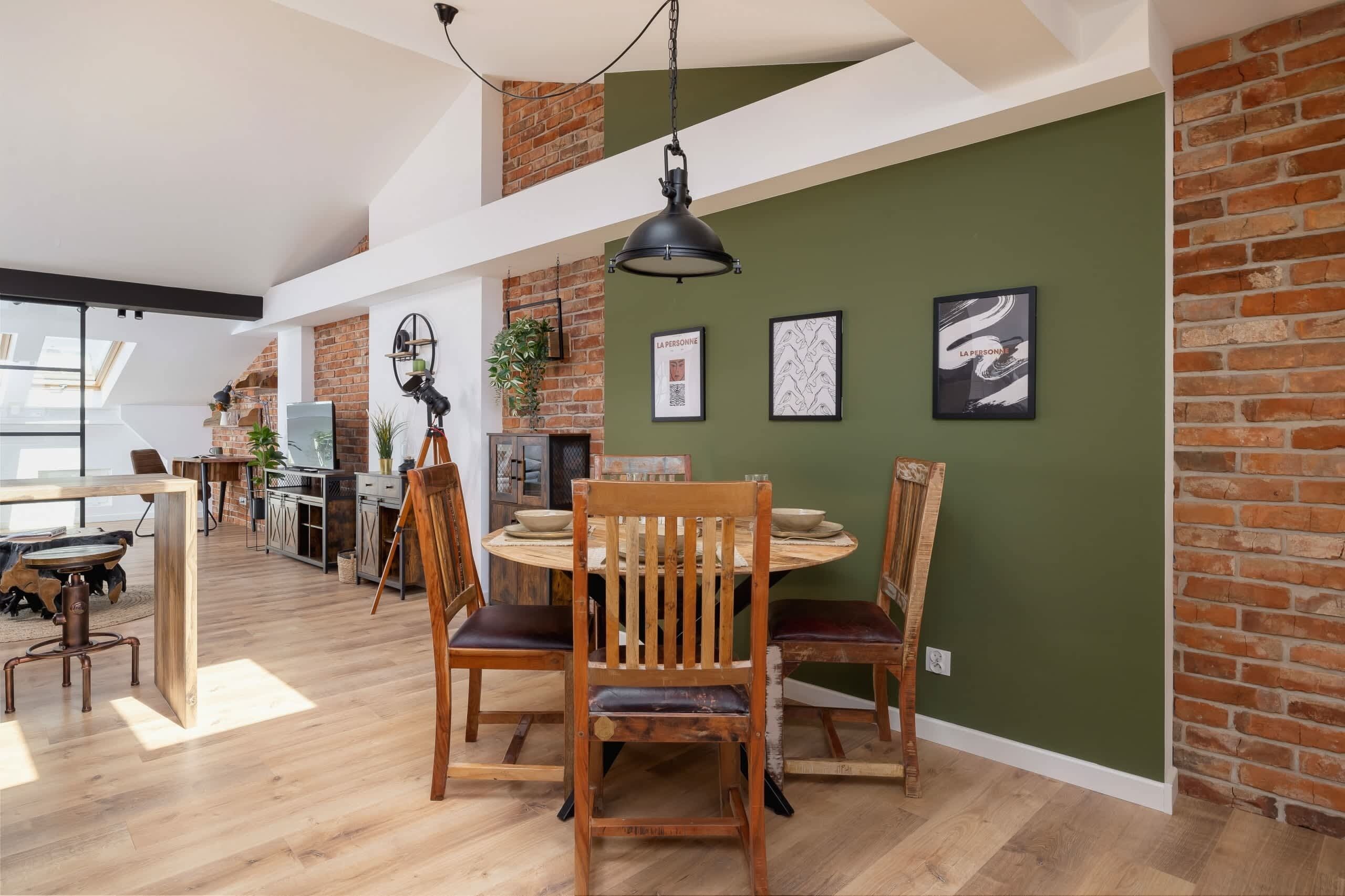 Un vaste espace dédié à la salle à manger, avec une table ronde en bois et des chaises, un luminaire suspendu de style industriel et un mur en briques orné d’œuvres d’art encadrées.