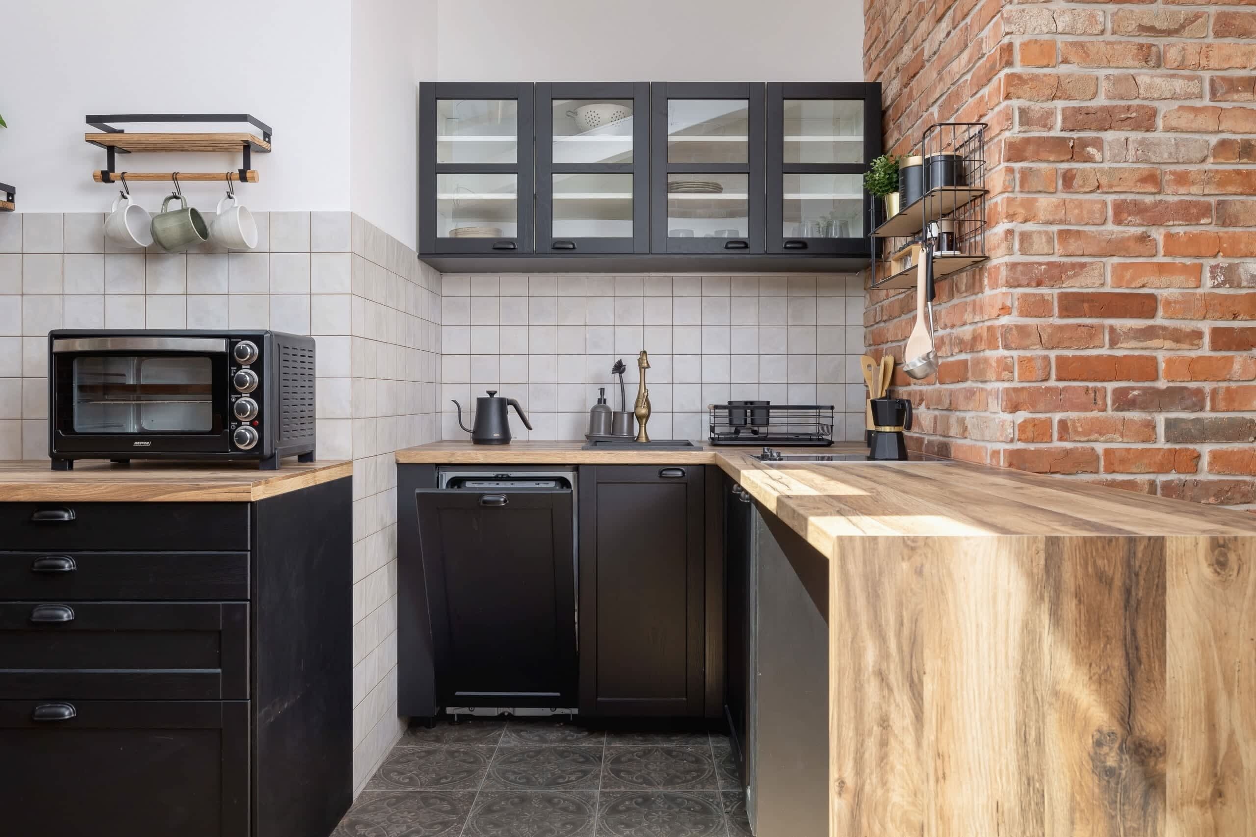 A compact kitchen with a built-in dishwasher, a microwave, and a coffee machine. There are cabinets with glass doors and a brick wall accent.
