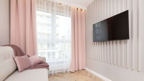 Ein modernes Wohnzimmer mit einer Couch, rosafarbenen Vorhängen, einem Wand-Fernseher und Blick auf die Stadt.
