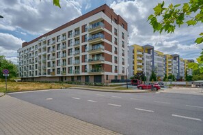 Um edifício residencial com vários andares e varandas, uma rua pavimentada e um carro vermelho estacionado perto.