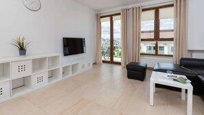 Una sala de estar moderna con un televisor de pantalla plana, una mesa de centro blanca, un sofá de cuero negro y un ventanal con cortinas.
