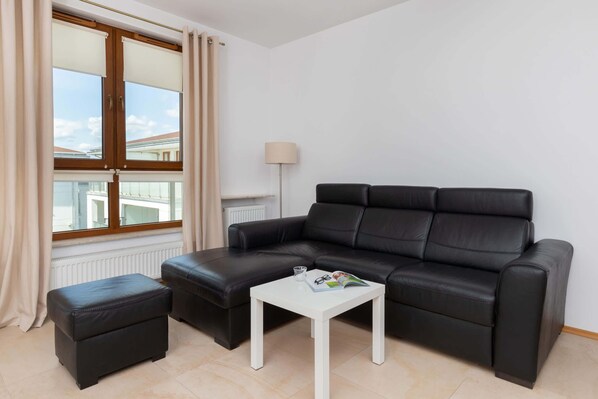 Una sala moderna con un sofá de cuero negro, una mesita de centro blanca y un pequeño otomaní. Se ve una ventana con cortinas, y una lámpara de pie está colocada contra la pared.
