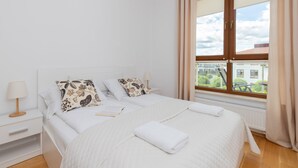 Un dormitorio con una cama grande, ropa de cama blanca, una mesita de noche con lámpara y una ventana con cortinas.