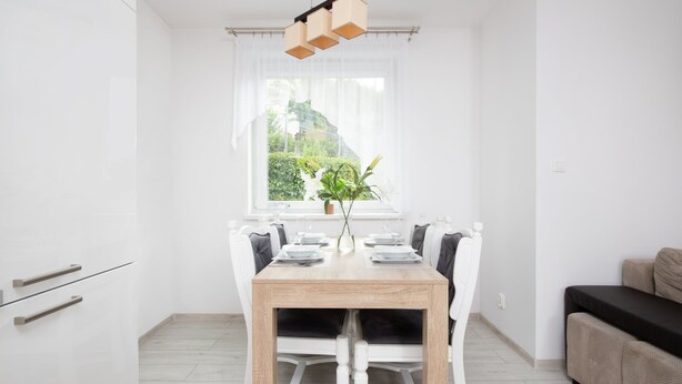 Un espace de repas lumineux et moderne, avec une table en bois et des chaises blanches, une fenêtre donnant sur un paysage verdoyant et un luminaire suspendu.