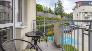 Un balcon avec une porte vitrée, une petite table ronde, une chaise en osier et une vue sur une piscine et des voitures garées.