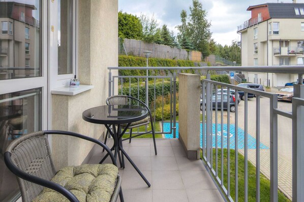 A balcony with a glass door, a small round table, a wicker chair, and a view of a swimming pool and parked cars.