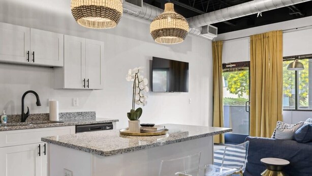 A modern kitchen with white cabinets, granite countertops, and a central island. There is a dining area with a blue sofa and clear chairs. - Modern Loft · Pet Friendly · 8 Min BNA · Fast WiFi (Nashville)