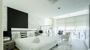 Une chambre moderne avec un grand lit, une table de chevet, une commode et une vue sur la ville grâce à de grandes fenêtres.