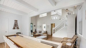 Une table de billard dans une pièce avec un escalier, des meubles en osier et un mur blanc orné d’une lettre « A ».