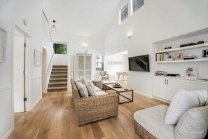 A spacious living area with a staircase, a wicker sofa, a white sofa, a coffee table, and a TV on a white cabinet.