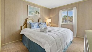 A bedroom with a large bed, a painting on the wall, a window with curtains, and a lamp. - Seafarer Unit 107 - Oceanview (North Myrtle Beach)