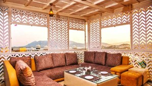 A modern, well-lit room with a large sectional sofa, a glass coffee table, and a view of distant mountains through large windows with patterned screens. - Pax Rafahiya Haveli Udaipur (Udaipur)