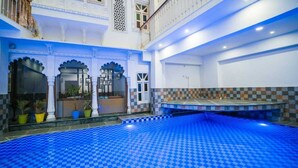 An indoor swimming pool with a blue checkered floor, surrounded by white walls with decorative arches and a balcony with railings. - Pax Rafahiya Haveli Udaipur (Udaipur)