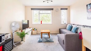 Un salon avec un canapé gris, une table basse en bois, un téléviseur, un ventilateur, une petite table avec une plante et une fenêtre donnant sur des arbres.