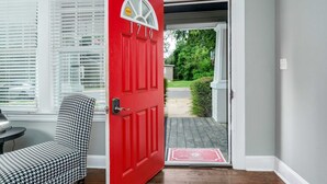 Una puerta roja con el número 1710, un sillón estampado con diseño pied de poule y un ventanal con marco blanco y persianas.