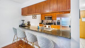 Una cocina con armarios de madera, encimera de granito, refrigerador de acero inoxidable y una zona de bar con taburetes.