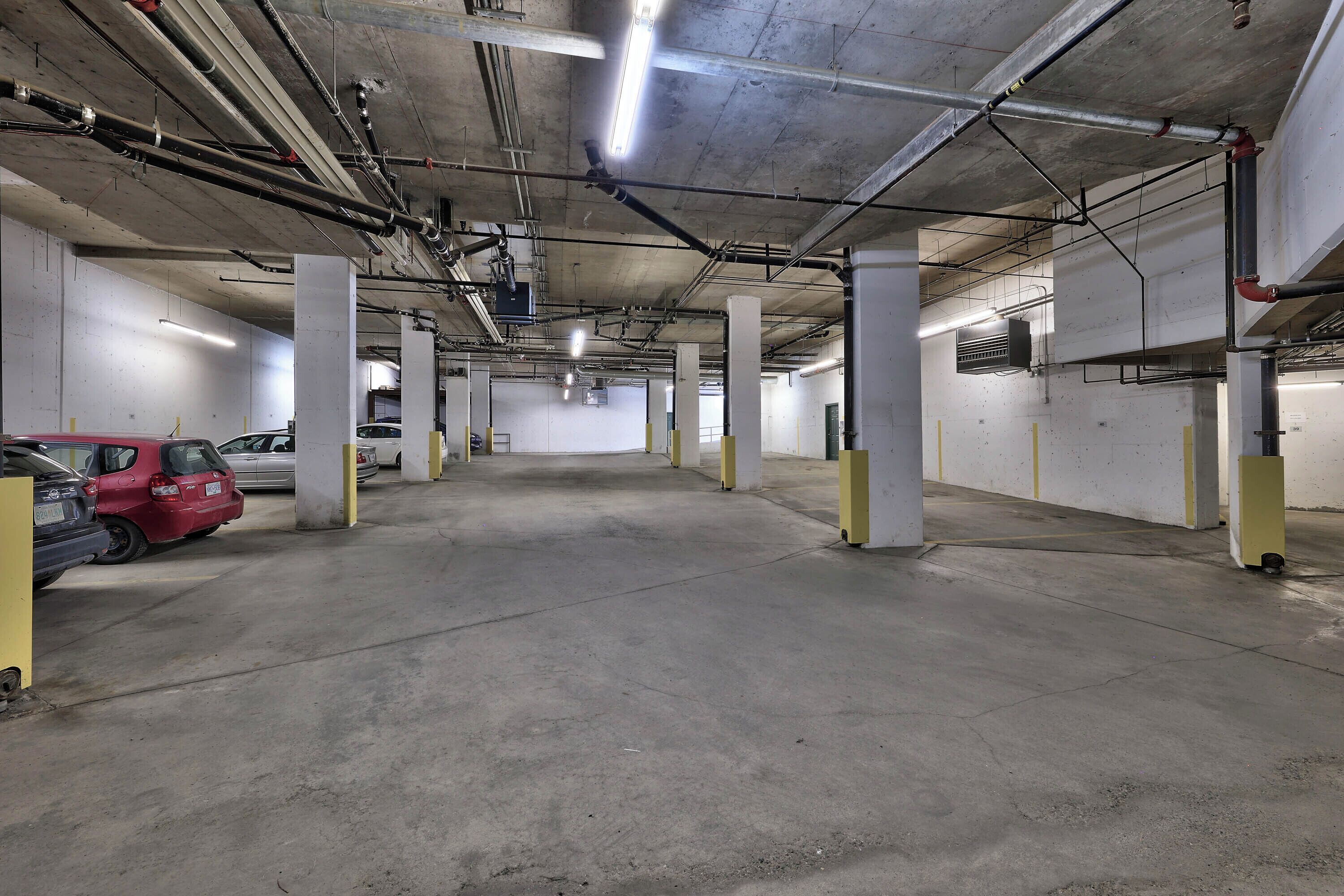 Un parking souterrain avec des planchers et des piliers en béton, un éclairage fluorescent et des tuyaux et des conduits apparents.