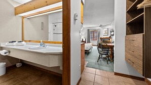 A bathroom with a large mirror, a sink, and a shower. There is a wooden shelf with towels and a chair in the adjacent room.