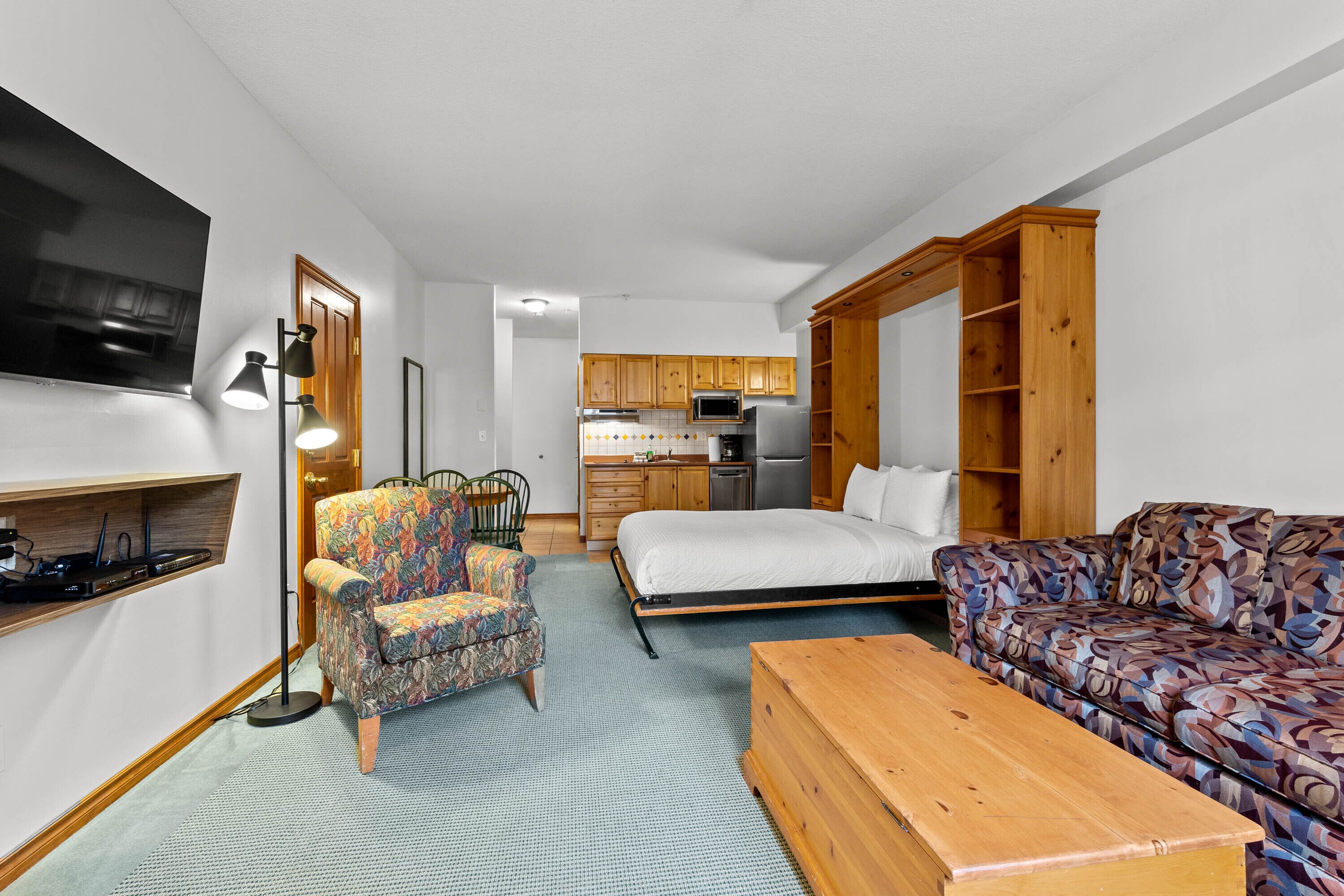 Un espace de vie compact comprenant un lit, un canapé, un fauteuil, une kitchenette et un téléviseur à écran plat fixé au mur.
