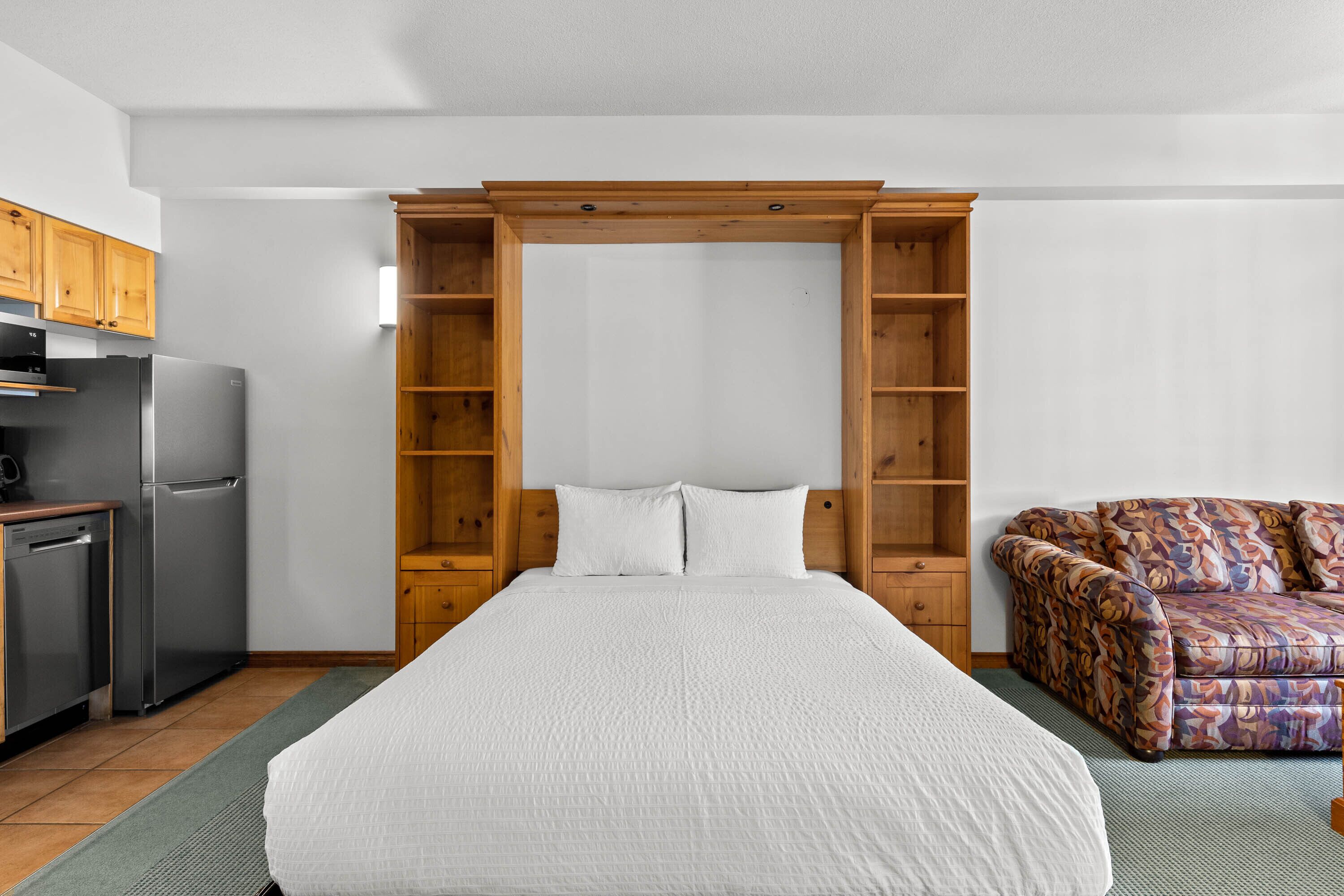 A compact room with a bed, a sofa, a kitchenette, and a wooden cabinet structure.