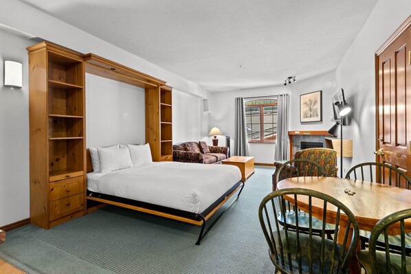 A bedroom with a bed, a wooden wardrobe, a sofa, a dining table with chairs, and a lamp.
