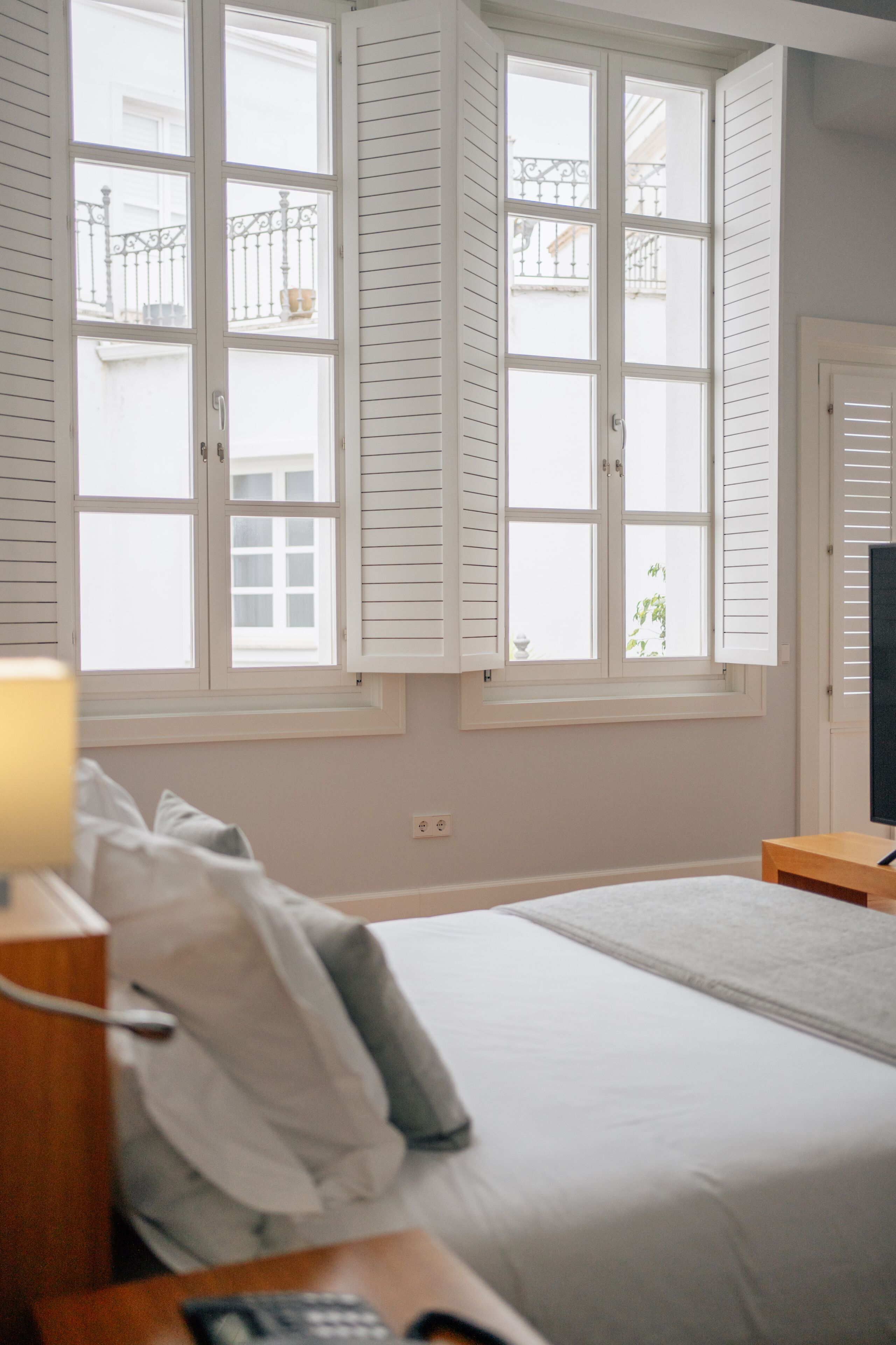 Un dormitorio con una cama, una mesita de noche, una lámpara y una ventana con contraventanas.