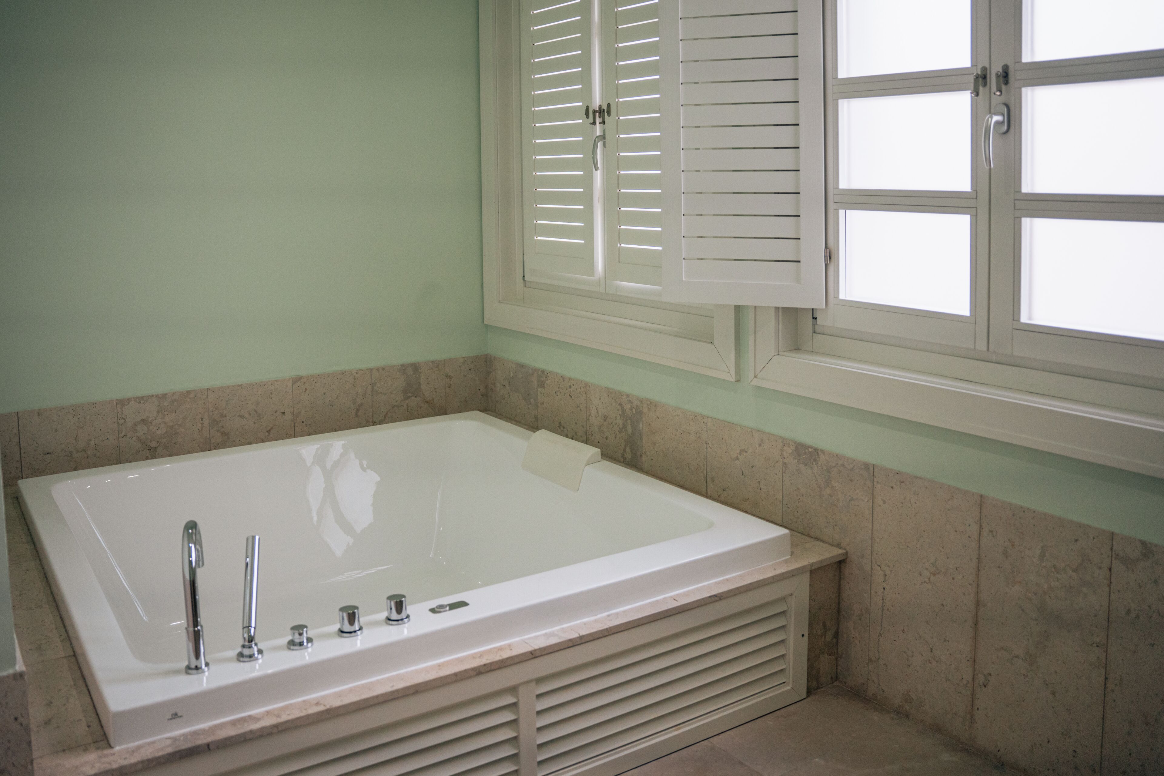 A bathroom with a large bathtub, a window with shutters, and a tiled wall.