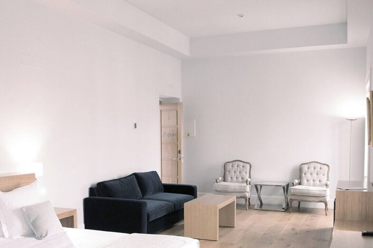 A modern hotel room with a bed, a dark blue sofa, two armchairs, a small table, and a desk.