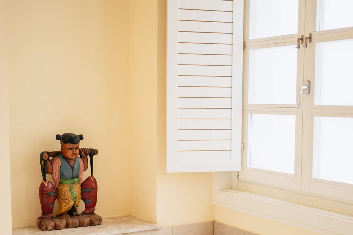 A bathroom with a bathtub, a window with shutters, and a shelf with a statue.
