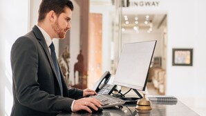 Een man in een pak achter een receptiebalie, met een laptop, telefoon en een kleine bel.