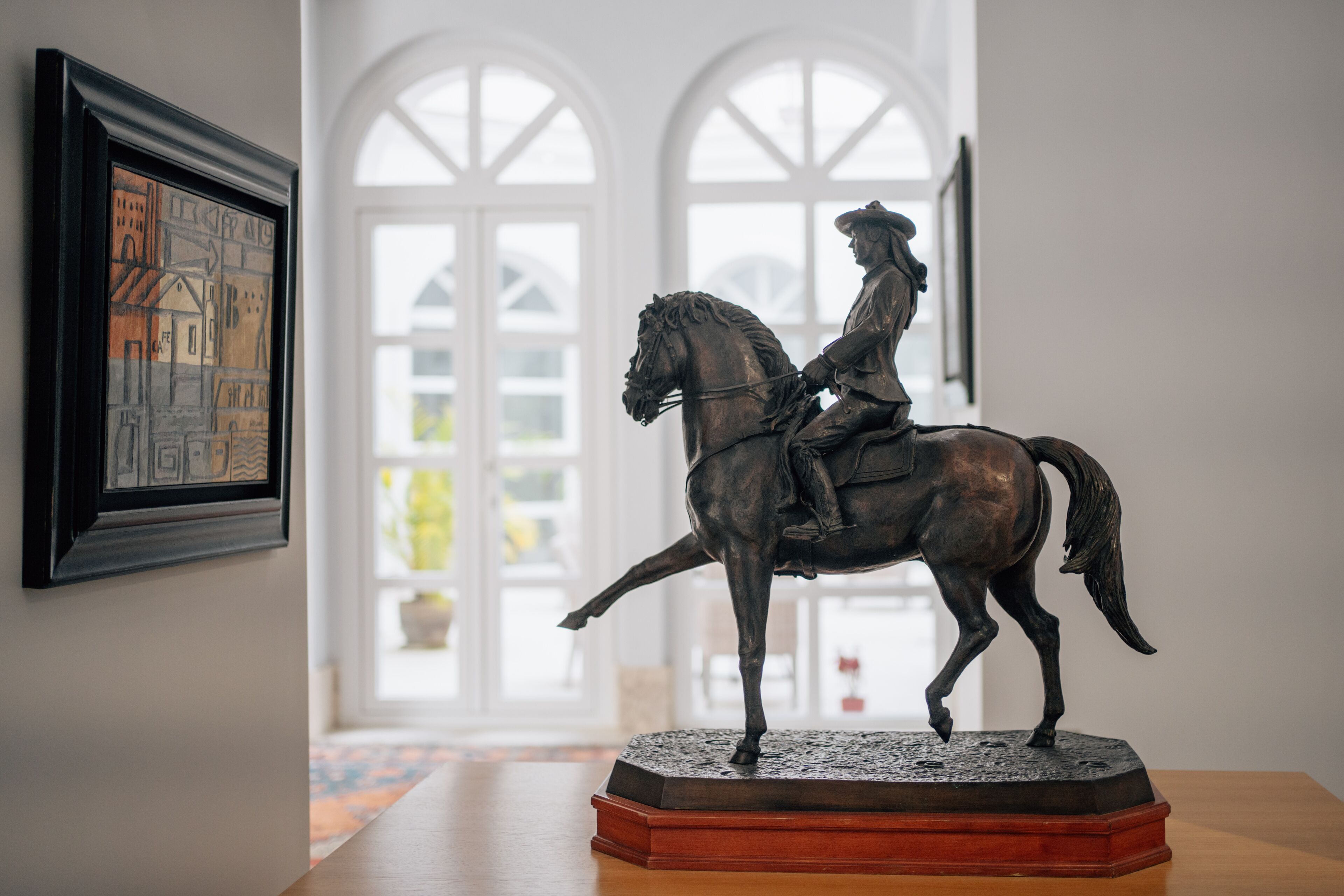 A statue of a man on a horse on a pedestal in a room with a large window and a framed picture on the wall.