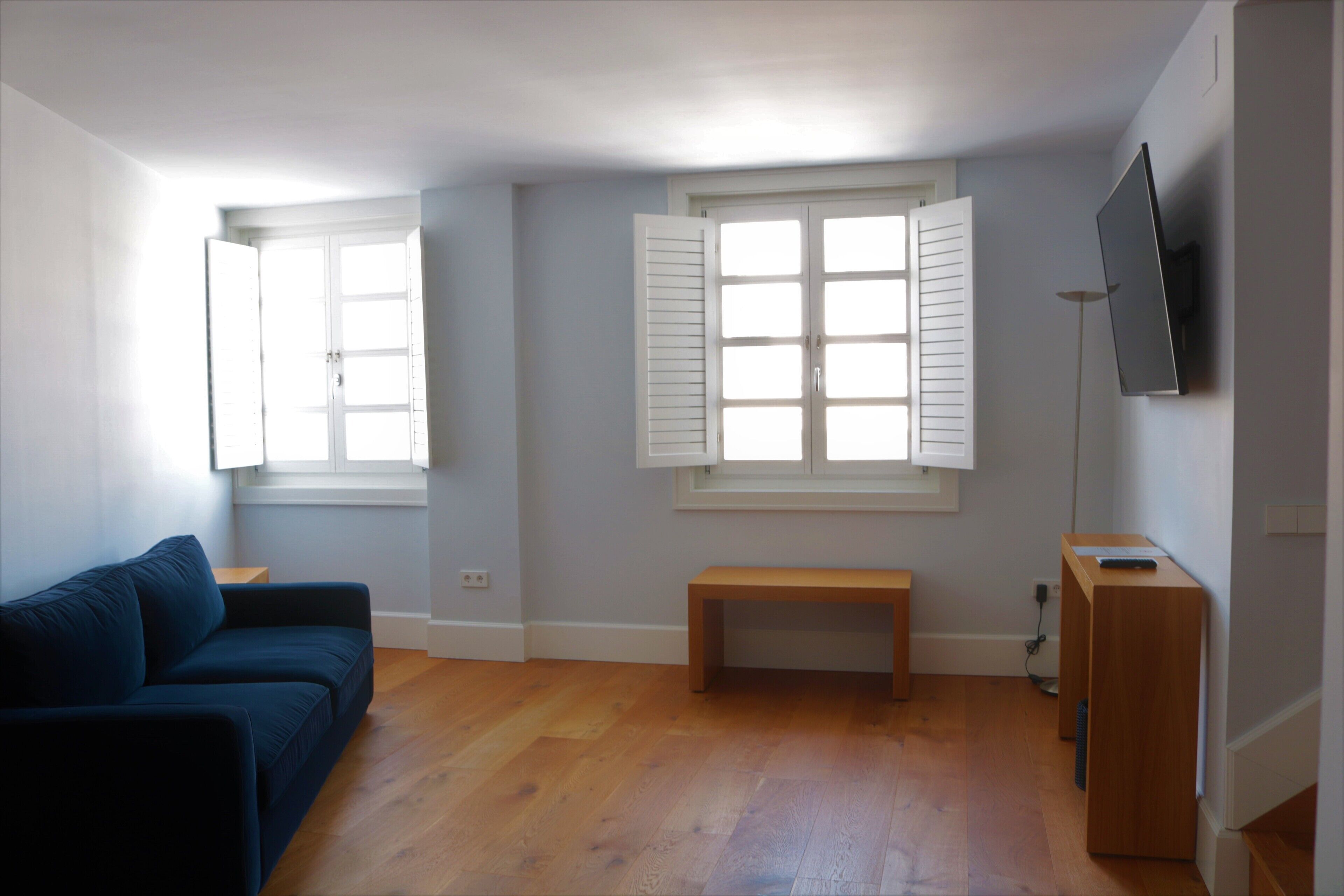 Ein Zimmer mit einem blauen Sofa, einer Holzbank, einem an der Wand montierten Fernseher und einem Fenster mit Rollläden.